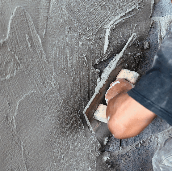 Application Enduit Mural : Pose de Ciment par un Maçon Main d'ouvrier appliquant du ciment gris texturé sur un mur avec une truelle. Surface d'enduit frais et rugueux en gros plan.