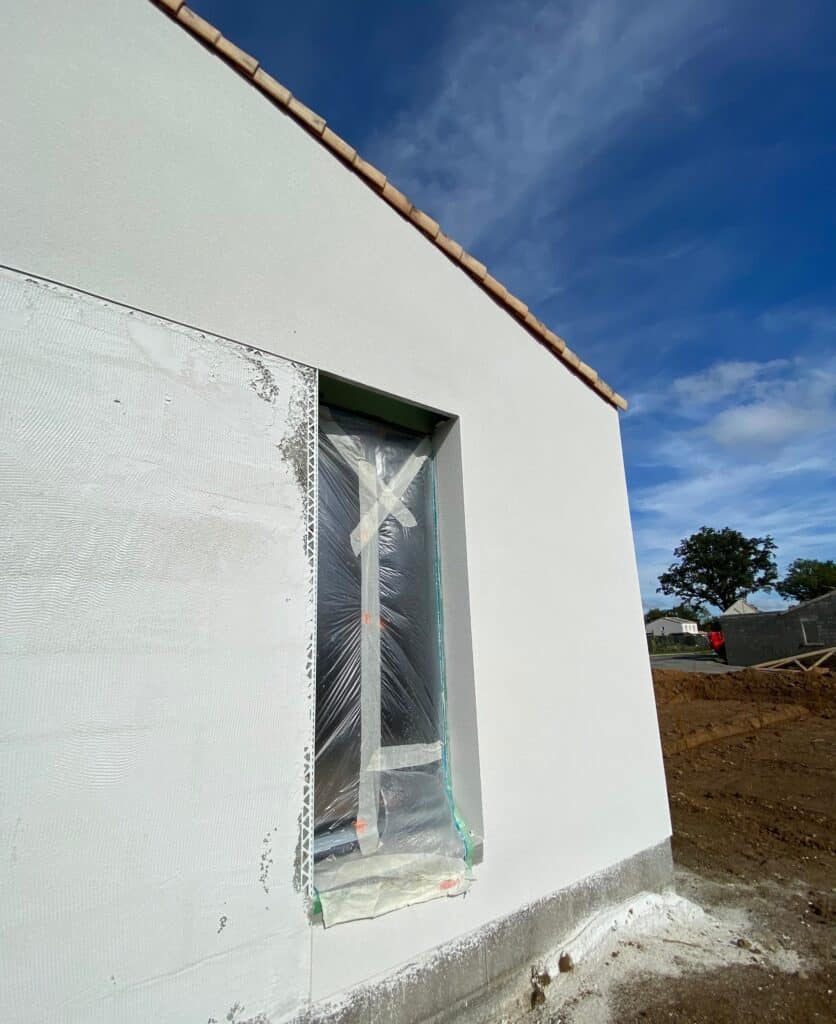 Façade Maison Neuve: Enduit Blanc et Fenêtre Bâchée Façade de maison en construction avec enduit blanc texturé, fenêtre bâchée, tuiles de toit et terre de chantier. Ciel bleu.