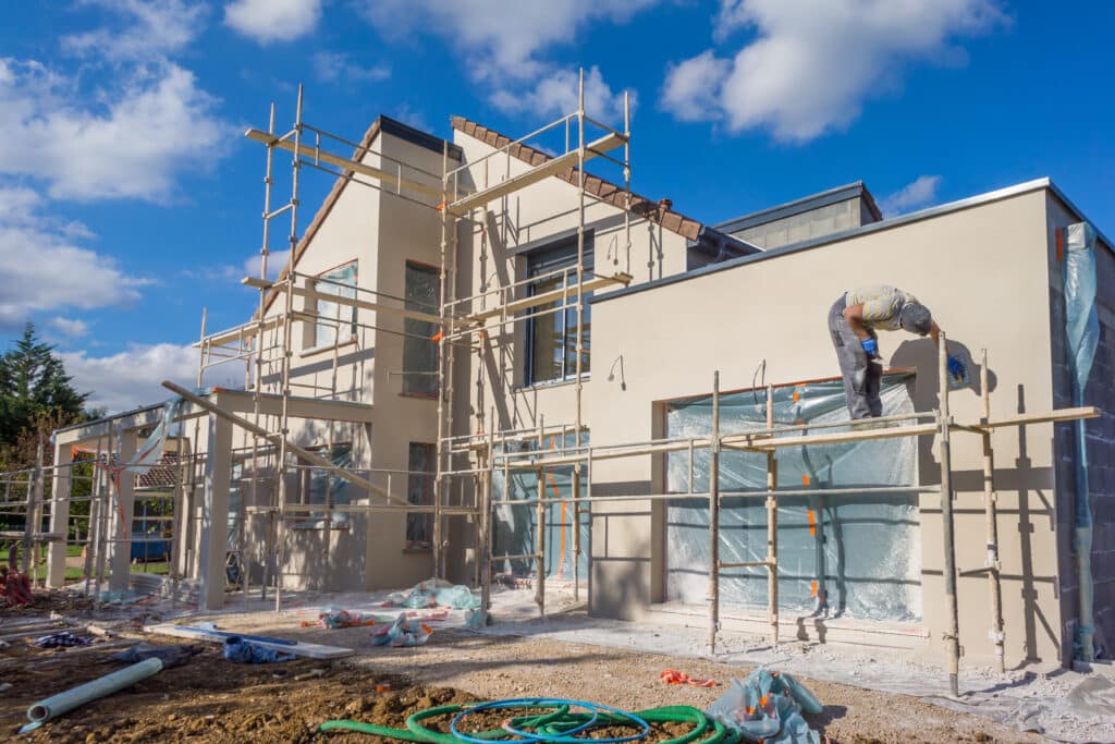 Ravalement maison : Ouvrier sur chantier de construction. Ouvrier sur échafaudage appliquant le ravalement sur la façade beige d'une maison en construction, sous ciel bleu.