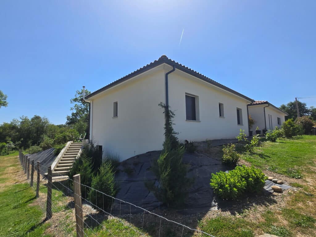 Maison Moderne et Aménagement de Jardin en Pente Maison contemporaine blanche sur terrain en pente avec escalier extérieur et jardin paysager sous ciel bleu.